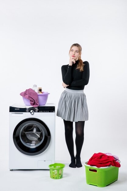 Vista frontal de la mujer joven con lavadora y ropa sucia pensando en la pared blanca