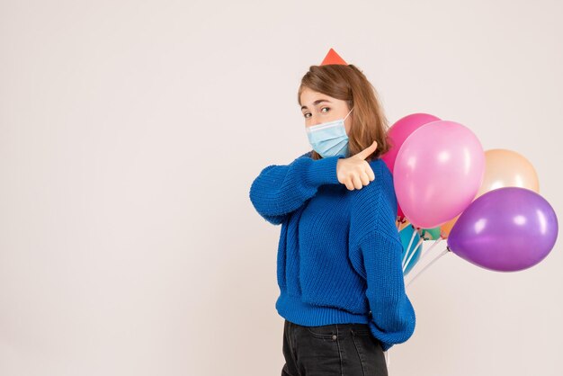 Vista frontal mujer joven en máscara estéril escondiendo globos de colores detrás de su espalda