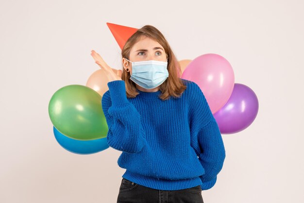 Vista frontal mujer joven en máscara estéril escondiendo globos de colores detrás de su espalda