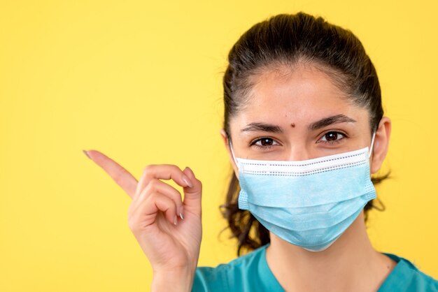 Vista frontal de la mujer joven con máscara médica apuntando con el dedo algo en la pared amarilla