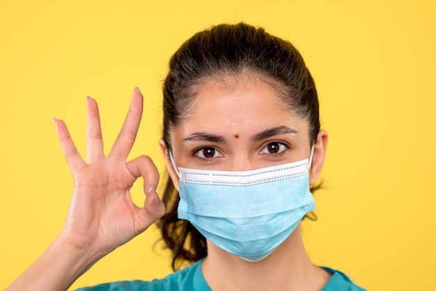 Vista frontal de la mujer joven con máscara médica haciendo okey firmar en la pared amarilla