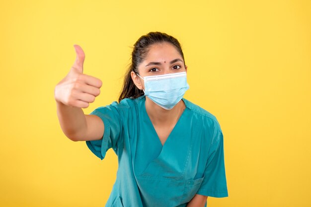 Vista frontal de la mujer joven con máscara médica haciendo pulgar hacia arriba firmar en la pared amarilla