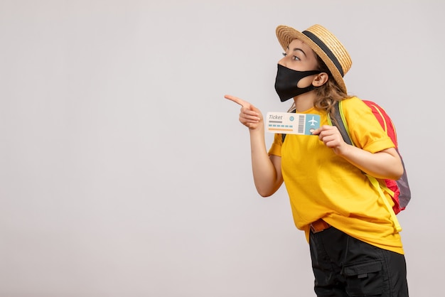 Vista frontal mujer joven con máscara negra con billete de viaje apuntando con el dedo a la izquierda