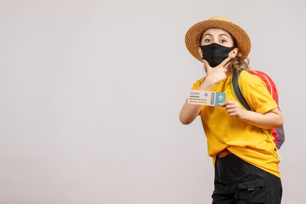 Vista frontal mujer joven con máscara negra con billete de viaje poniendo la mano en la barbilla
