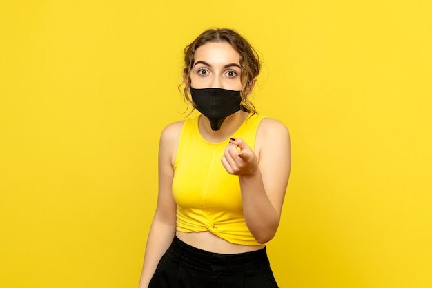 Vista frontal de la mujer joven en máscara negra sorprendida en la pared amarilla
