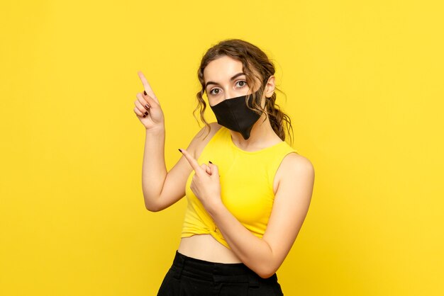 Vista frontal de la mujer joven con máscara en la pared de color amarillo claro