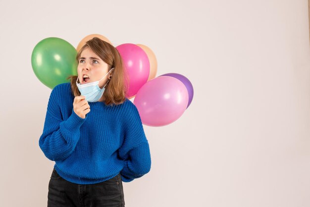 Vista frontal mujer joven en máscara sosteniendo globos de colores detrás de su espalda