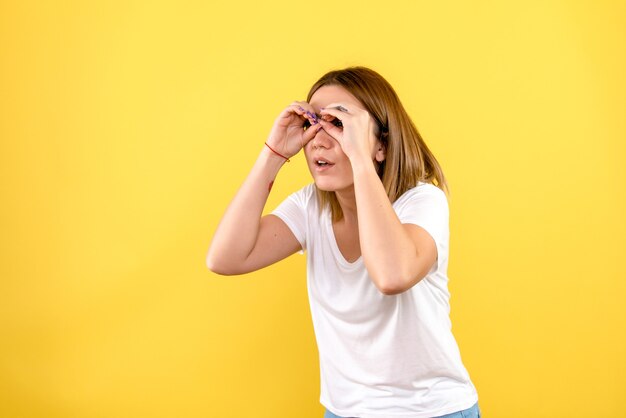 Vista frontal de la mujer joven mirando a través de los dedos en la pared amarilla