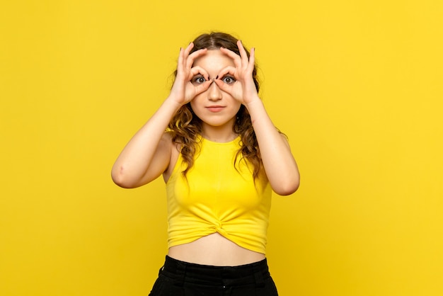 Vista frontal de la mujer joven mirando a través de los dedos en la pared amarilla
