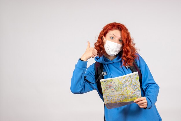 Vista frontal de la mujer joven con mochila y mapa en máscara sobre una pared blanca