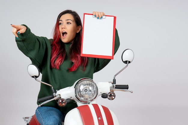 Vista frontal mujer joven en motocicleta sosteniendo nota para firma apuntando en la pared blanca