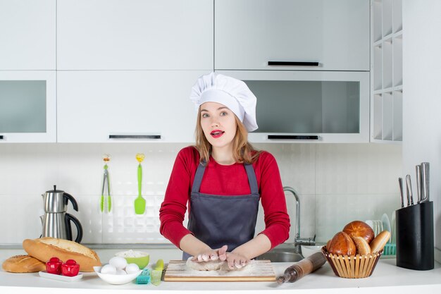 Vista frontal mujer joven ocupada en sombrero de cocinero y delantal amasando masa en la cocina