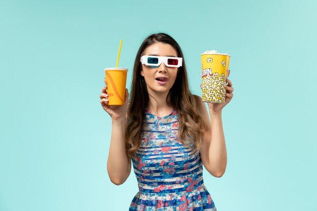 Vista frontal mujer joven con paquete de palomitas de maíz y bebida en gafas de sol d sobre superficie azul claro