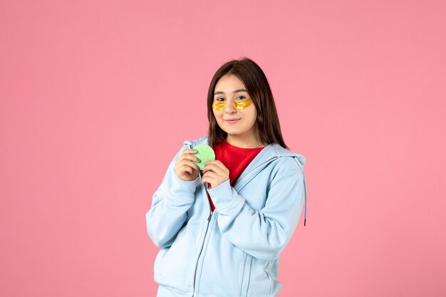 Vista frontal de la mujer joven con parches en los ojos y una pequeña esponja en la pared rosa