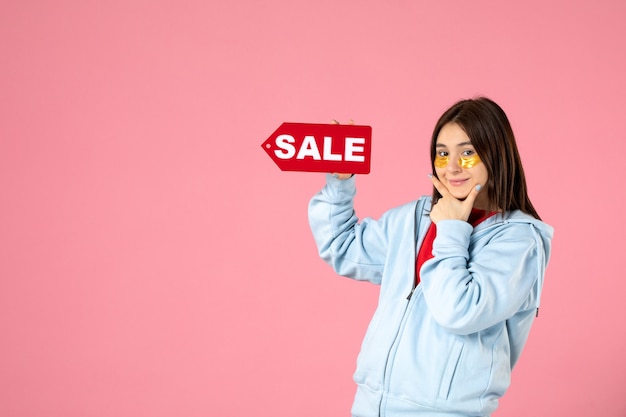 Vista frontal de la mujer joven con parches en los ojos sosteniendo un banner de venta rojo en la pared rosa