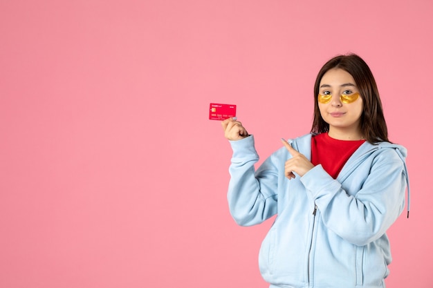 Vista frontal de la mujer joven con parches en los ojos sosteniendo una tarjeta bancaria en la pared rosa