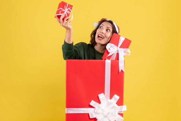 Vista frontal de la mujer joven de pie dentro de la caja con regalos de Navidad en la pared amarilla
