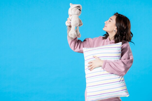 Vista frontal mujer joven en pijama rosa con oso de juguete y almohada en azul