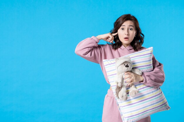 Vista frontal mujer joven en pijama rosa con oso de juguete y almohada en azul