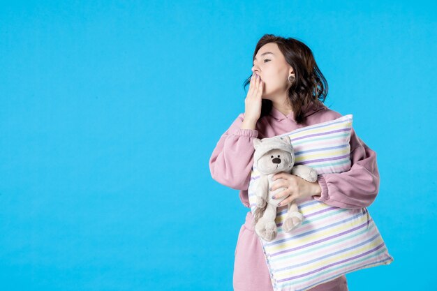 Vista frontal mujer joven en pijama rosa con oso de juguete y almohada bostezando en azul