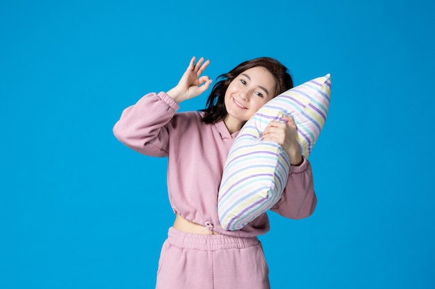 Vista frontal mujer joven en pijama rosa sosteniendo la almohada en la pared azul noche color fiesta insomnio reposo en cama sueño mujer emoción