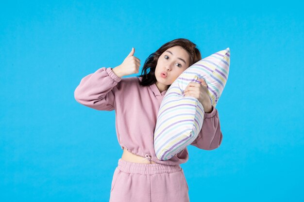 Vista frontal mujer joven en pijama rosa sosteniendo la almohada en la pared azul noche color fiesta insomnio reposo en cama sueño mujer emociones dormir