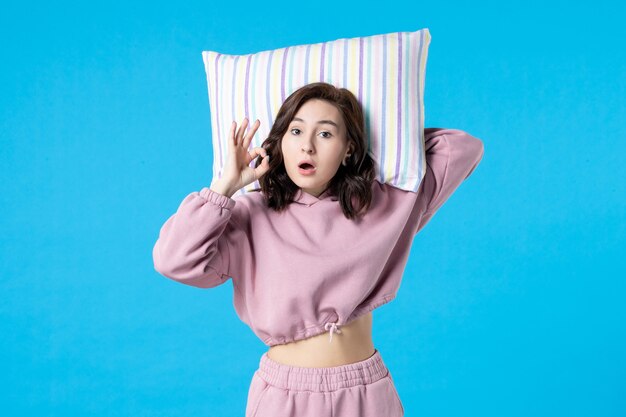 Vista frontal mujer joven en pijama rosa sosteniendo la almohada en la pared azul noche color insomnio dormir reposo en cama sueño mujer fiesta emoción