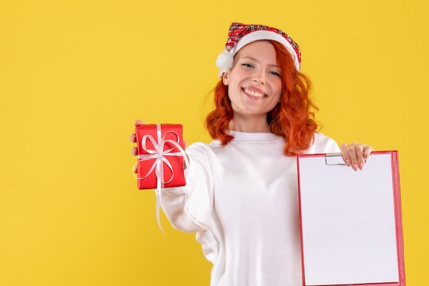 Vista frontal de la mujer joven con poco presente de Navidad y nota de archivo en la pared amarilla