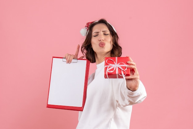 Vista frontal de la mujer joven con poco presente de Navidad y nota de archivo en la pared rosa