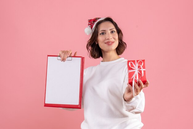 Vista frontal de la mujer joven con poco presente de Navidad y nota de archivo en la pared rosa