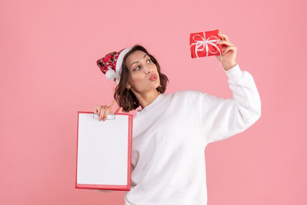 Vista frontal de la mujer joven con poco presente de Navidad y nota de archivo en la pared rosa