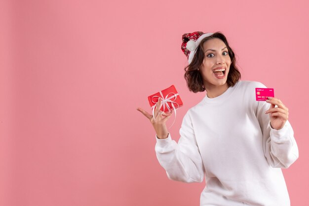 Vista frontal de la mujer joven con poco presente de Navidad y tarjeta bancaria en la pared rosa