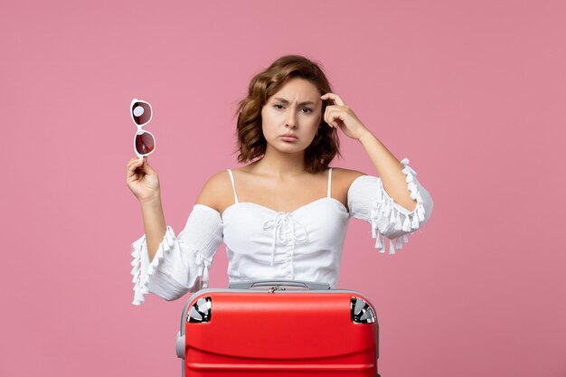 Vista frontal de la mujer joven posando y preparándose para el viaje con bolsa roja en la pared rosa