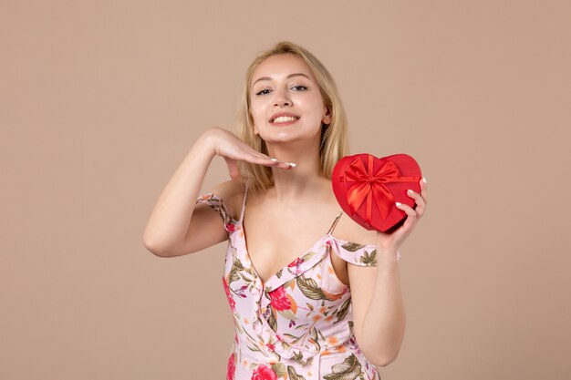 Vista frontal de la mujer joven posando con presente en forma de corazón rojo en la pared marrón