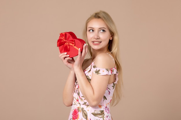 Vista frontal de la mujer joven posando con presente en forma de corazón rojo en la pared marrón