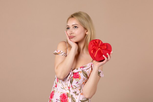 Vista frontal de la mujer joven posando con presente en forma de corazón rojo en la pared marrón