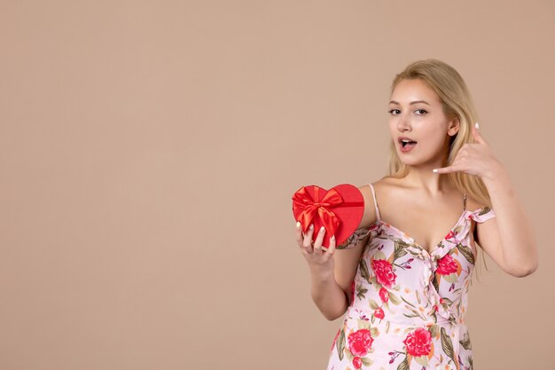 Vista frontal de la mujer joven posando con presente en forma de corazón rojo en la pared marrón