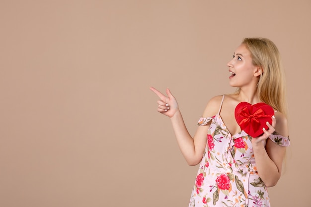 Vista frontal de la mujer joven posando con presente en forma de corazón rojo en la pared marrón