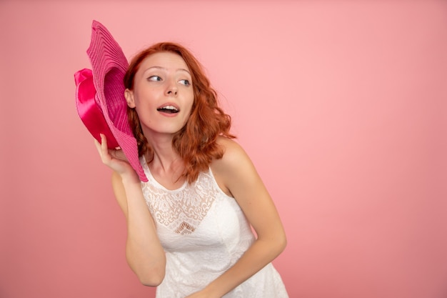 Vista frontal de la mujer joven posando con sombrero rosa en la pared rosa