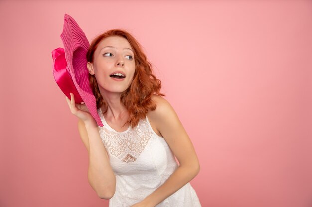Vista frontal de la mujer joven posando con sombrero rosa en la pared rosa