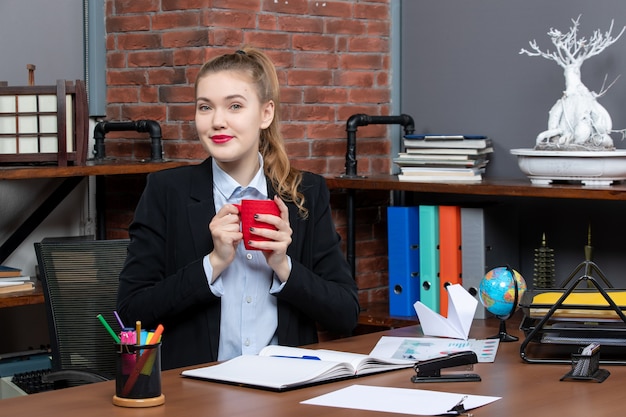 Vista frontal de la mujer joven positiva sentada en una mesa y sosteniendo una taza roja en la oficina