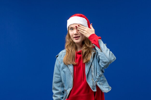 Vista frontal mujer joven que cubre la mitad de su rostro sobre fondo azul color de emociones navideñas