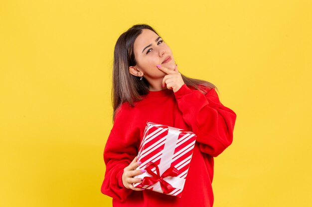 Vista frontal de la mujer joven con regalo de Navidad pensando en la pared amarilla