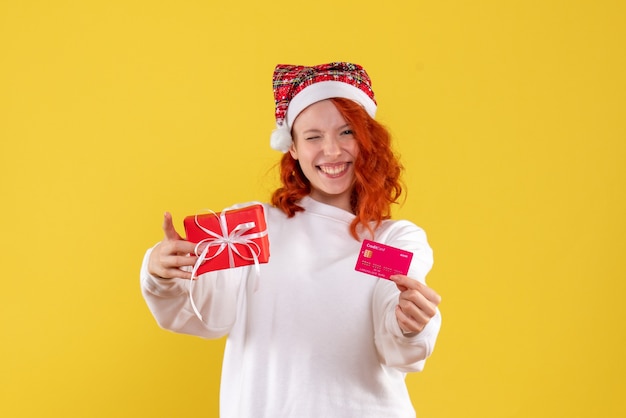 Vista frontal de la mujer joven con regalo de Navidad y tarjeta bancaria en la pared amarilla
