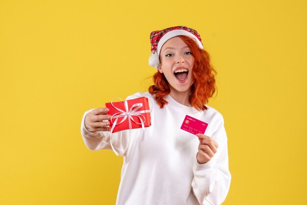 Vista frontal de la mujer joven con regalo de Navidad y tarjeta bancaria en la pared amarilla