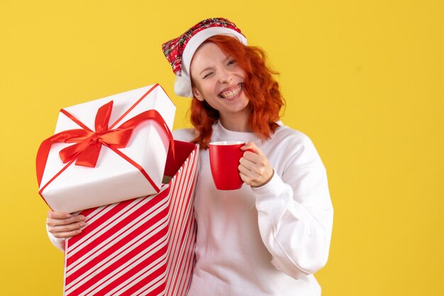 Vista frontal de la mujer joven con regalo de Navidad y una taza de té en la pared amarilla