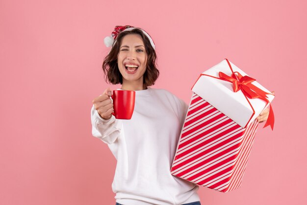 Vista frontal de la mujer joven con regalo de Navidad y una taza de té en la pared rosa