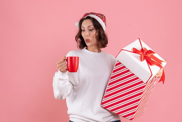 Vista frontal de la mujer joven con regalo de Navidad y una taza de té en la pared rosa