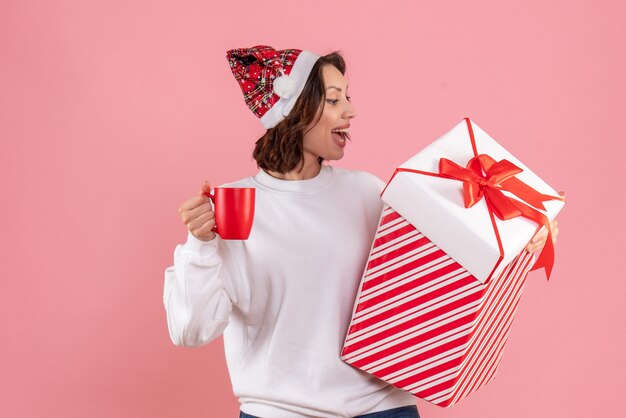 Vista frontal de la mujer joven con regalo de Navidad y una taza de té en la pared rosa