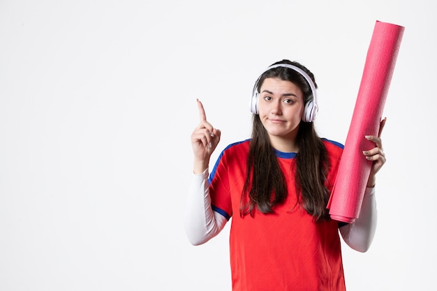 Vista frontal mujer joven en ropa deportiva con estera de yoga escuchando música pared blanca